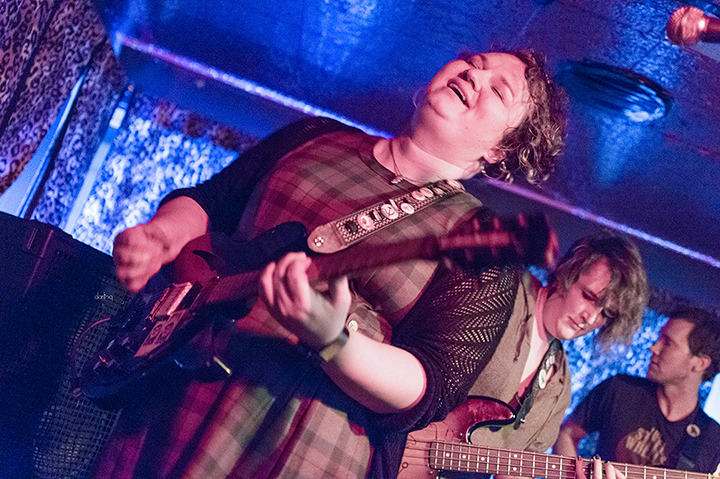 A photo of Josie Cool playing guitar and performing as part of Teacup Gorilla. 2 other band members are shown behind her