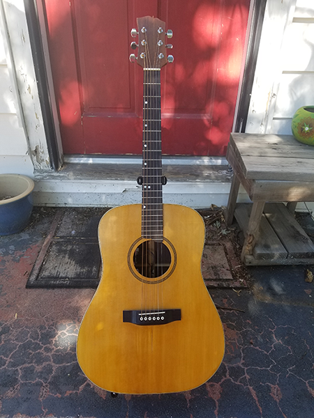 An image of Josselyn Cool Guitar Works 001, a pre-war Martin D18 style acoustic guitar. It's siting on a cement porch in front of a red door.